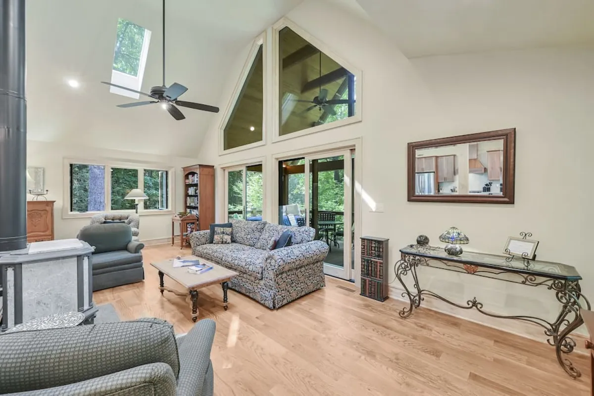 The hero image showcases a warm and inviting living room with oversized windows framing woodland views, a plush sofa gathered around a crackling fireplace, a bookshelf brimming with reads, and a coffee table perfect for morning brews or evening conversation. Soft lighting and cozy décor create an atmosphere ideal for relaxation and togetherness.