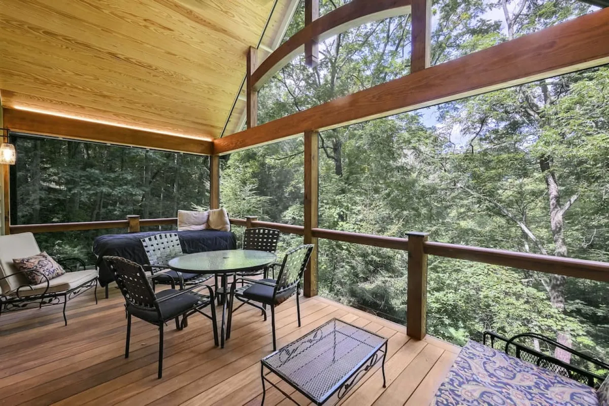 This exterior shot features a spacious outdoor seating area with a dining table, wooden beams, and large windows looking into the home. Surrounded by green trees, the area invites relaxation, al fresco meals, and sunset watching, all set in a peaceful forest backdrop.