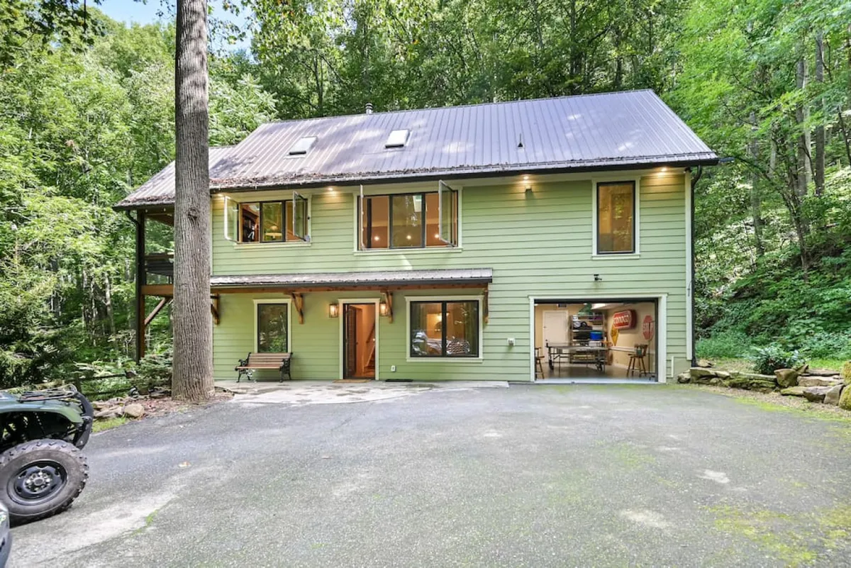 The green exterior of the house is nestled among tall trees, with large windows and a driveway leading up to it. The setting is welcoming and tranquil, offering glimpses of the surrounding forest and an inviting entry point to the estate.