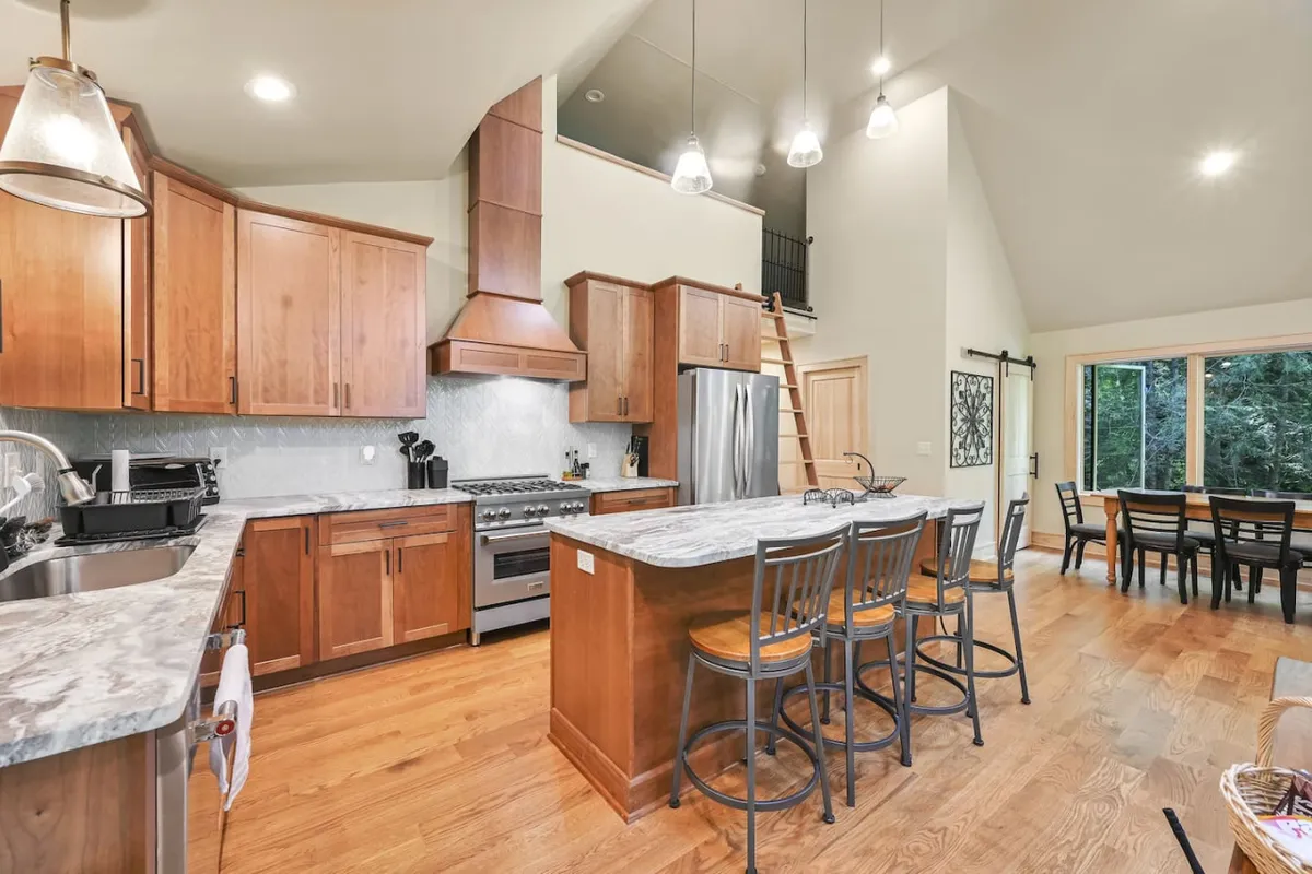 A bright and modern kitchen space with a prominent island, stainless steel appliances, large windows flooding the room with natural light, and warm wooden cabinetry. The scene captures the energy of family gatherings and the pleasures of both casual and celebratory meals.