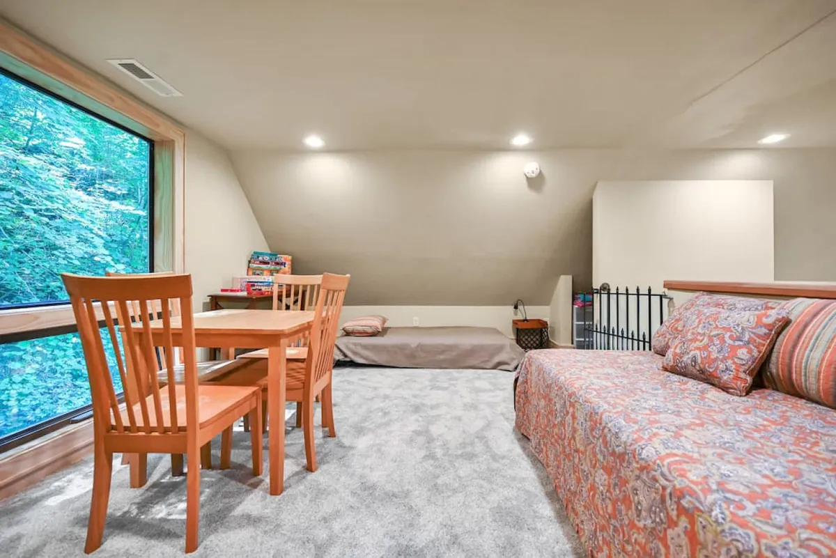 A cozy lounge area with a sofa bed adorned with decorative pillows sits beside a dining table and chairs, all facing a large window that offers a breathtaking forest vista. The natural light and warm interior palette create an inviting space for relaxation, board games, or meals with loved ones.