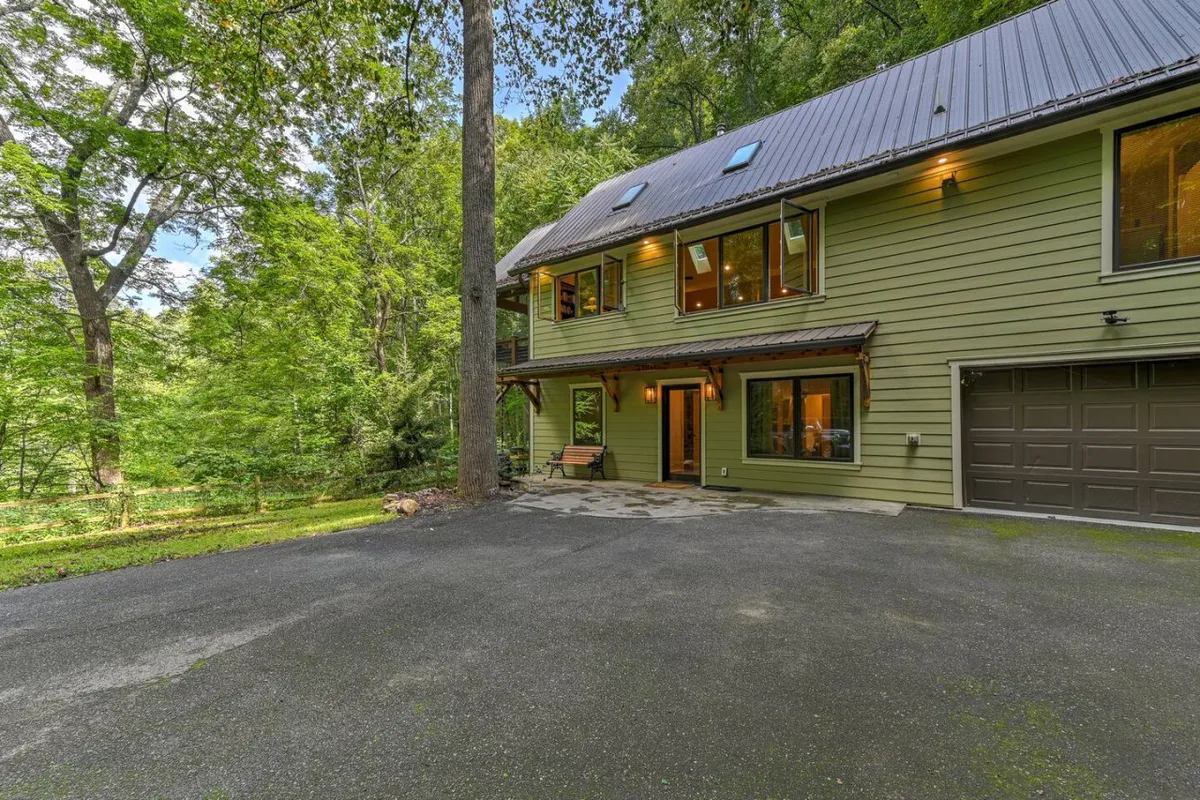 This image captures the modern architecture of the property, featuring large windows that hint at the connection to nature, a welcoming covered porch, a driveway, and a lush border of surrounding trees. The scene is serene and welcoming, with a sense of seclusion while remaining inviting for guests to return after a day out.