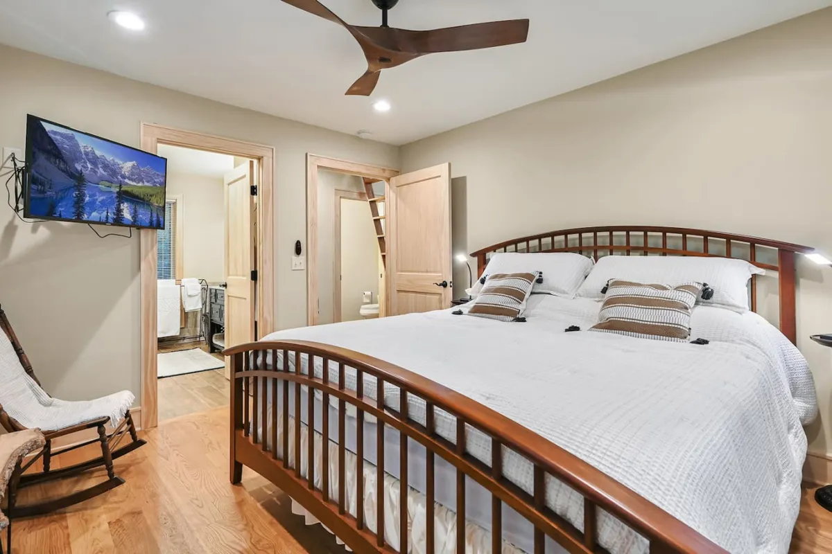 An inviting bedroom with a comfortable bed, a TV placed for in-bed viewing, a classic wooden rocking chair, and a wooden door. Soft natural light filters in, enhancing the warm, restful ambiance ideal for both families and couples.