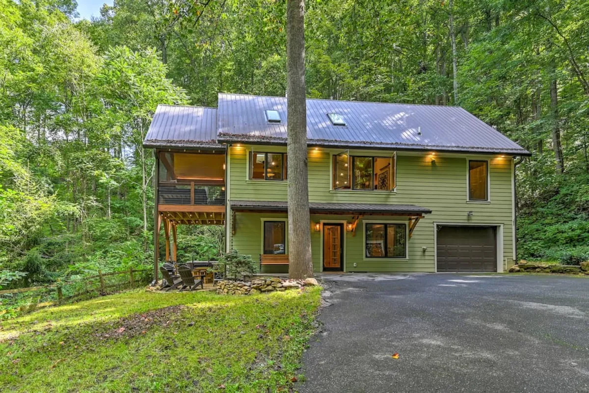 This image features the main house surrounded by trees, with large windows and a wooden deck. The setting is peaceful and inviting, with ample outdoor space for family activities and relaxation.