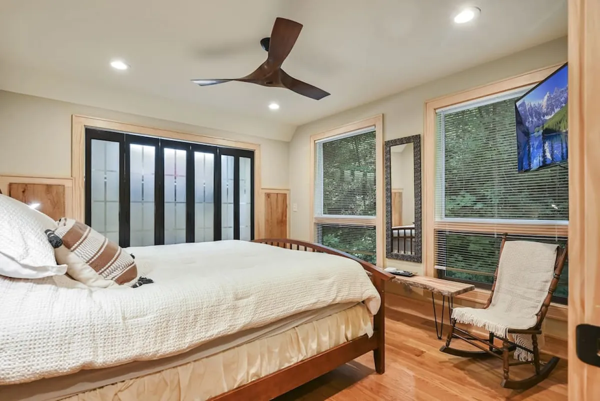 A serene bedroom featuring a plush bed positioned near large windows, which invite natural light and offer glimpses of the surrounding nature. The ceiling fan, mirror, and television add comfort and convenience, creating an inviting atmosphere perfect for relaxation, reading, or watching TV.